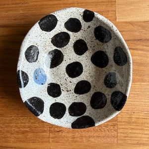 May include: A small, round, ceramic bowl with an irregular, hand-formed edge. The surface is off-white with a speckled texture and features numerous hand-painted, solid black circular spots of varying sizes, plus one distinct light blue spot.
