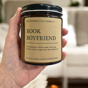May include: A soy candle in a clear glass jar with a black lid. The label reads "BOOK BOYFRIEND" and lists the scent notes: fresh mahogany, blood orange, clary sage, bourbon vanilla, tonka bean, and sandalwood. The candle is 9 oz.