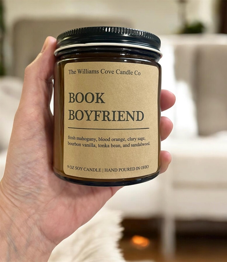 May include: A soy candle in a clear glass jar with a black lid. The label reads "BOOK BOYFRIEND" and lists the scent notes: fresh mahogany, blood orange, clary sage, bourbon vanilla, tonka bean, and sandalwood. The candle is 9 oz.