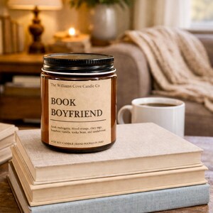 May include: A soy candle in a glass jar with a black lid, labeled "Book Boyfriend." The label includes scent notes and is placed on a stack of books. A white mug of coffee sits nearby, with a warm, cozy background.