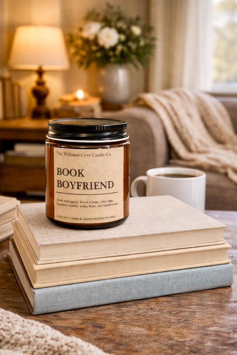 May include: A soy candle in a glass jar with a black lid, labeled "Book Boyfriend." The label includes scent notes and is placed on a stack of books. A white mug of coffee sits nearby, with a warm, cozy background.