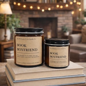 May include: Two soy candles with black lids and brown labels that read "BOOK BOYFRIEND." The candles sit on a stack of books. The background includes a fireplace and a string of lights.
