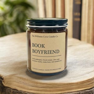 May include: A glass jar candle with a black lid and a label that reads "BOOK BOYFRIEND." The label lists the scent notes: fresh mahogany, blood orange, clary sage, bourbon vanilla, tonka bean, and sandalwood. The candle is 9 oz and hand-poured in Ohio.
