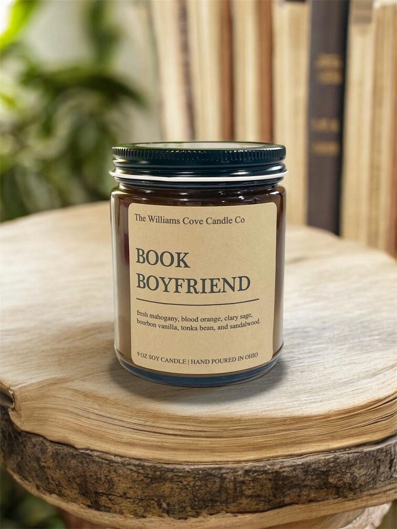May include: A glass jar candle with a black lid and a label that reads "BOOK BOYFRIEND." The label lists the scent notes: fresh mahogany, blood orange, clary sage, bourbon vanilla, tonka bean, and sandalwood. The candle is 9 oz and hand-poured in Ohio.