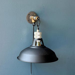 May include: Black industrial-style wall sconce with a matte finish. The lamp features a metal shade, a silver and brass bulb holder, and a wall-mounted fixture. The design incorporates pipe fittings and a chain for a vintage aesthetic.