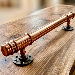 May include: A copper-colored industrial-style door pull or handle. The handle is made of copper pipes and fittings, with a diamond pattern etched into the central section. It is mounted on two black, circular flanges.