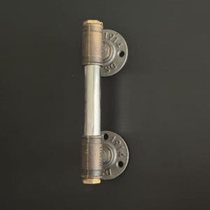 May include: Industrial-style toilet paper holder made from metal pipes and fittings. The holder features a silver roller and bronze-colored pipe sections, with circular mounting plates. The plates have the text "1914" and "DS" stamped on them.