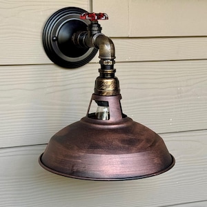 May include: A copper-colored industrial-style wall light fixture with a black metal base and a red valve. The light fixture has a round, copper-colored shade and a clear glass bulb.