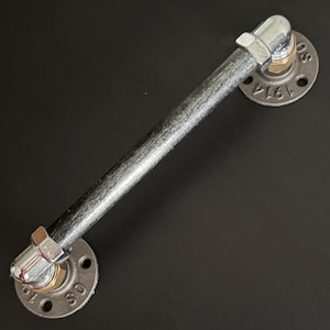 May include: A rustic, industrial-style door handle. The handle is made of a gray metal pipe with silver fittings and circular mounting plates. The plates have the number "1914" and the letters "SO" stamped on them. The handle is designed to be mounted horizontally.
