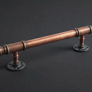 May include: A horizontal door handle constructed from a copper-toned pipe with two dark gray circular metal flanges at each end. The handle features a cylindrical design with visible joints and small diamond-shaped markings on the pipe surface.