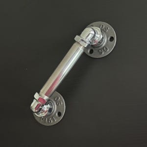 May include: A silver-colored industrial pipe handle with two circular flanges. The handle is made of a straight pipe section connected to angled fittings. The flanges have bolt holes and stamped text. The hardware is set against a dark background.