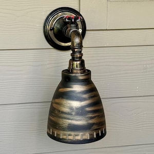 May include: A gold and black industrial style wall sconce with a pipe design and a bell-shaped shade. The sconce has a red valve handle.