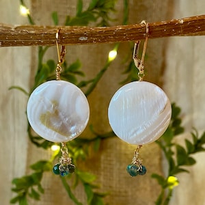 May include: A pair of white, round earrings with a pearlescent sheen. Each earring features a gold-colored leverback clasp and a cluster of small, green and blue beads. The earrings are suspended from a gold wire.