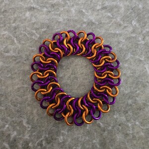 May include: A circular chainmail bracelet in a vibrant combination of purple and gold. The interlocking metal rings create a textured, decorative pattern. The bracelet is designed to be worn as a fashion accessory.