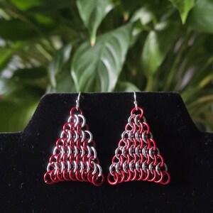 May include: A pair of handmade chainmail earrings. Each earring is a triangle shape, constructed with alternating red and silver metal rings. The earrings hang from silver-colored ear wires. The background is a blurred green.