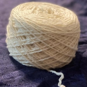 May include: A large, off-white ball of yarn sits on a dark blue, textured surface. The yarn has a slightly textured appearance, with a loose strand extending from the ball. Ideal for knitting and crochet projects.