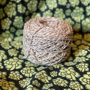May include: A ball of light brown yarn, tightly wound, sits on a black fabric patterned with green and yellow floral designs. The yarn appears to be a natural fiber, suitable for knitting or crochet projects.