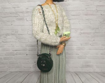 May include: A doll wearing a light green and white striped skirt, a light gray sweater, a gold necklace with green leaf charms, and green leaf earrings. The doll is holding a green phone and has a black round purse with a silver chain strap.