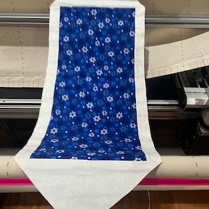 May include: A white fabric table runner with a blue fabric insert featuring a pattern of white Stars of David on a blue background. The runner is being prepared for sewing on a machine.