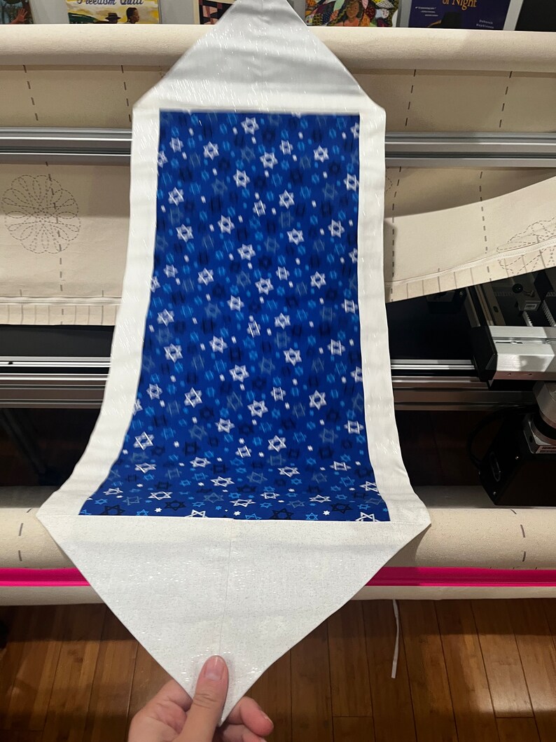 May include: A white fabric table runner with a blue fabric insert featuring a pattern of white Stars of David on a blue background. The runner is being prepared for sewing on a machine.