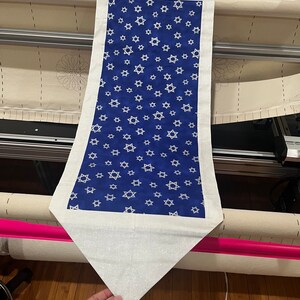May include: A white table runner with a blue centre panel featuring a repeating pattern of white Stars of David. The runner has a white border and a pointed end.