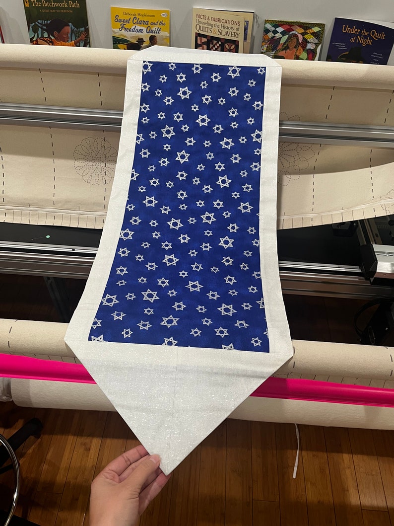 May include: A white table runner with a blue centre panel featuring a repeating pattern of white Stars of David. The runner has a white border and a pointed end.
