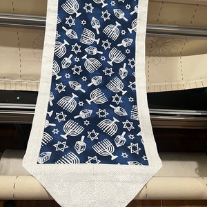 May include: A white fabric table runner with a blue background and a repeating pattern of white menorahs and stars. The runner has a white border and a pointed end.