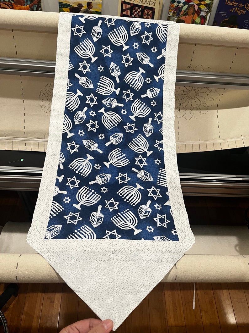 May include: A white fabric table runner with a blue background and a repeating pattern of white menorahs and stars. The runner has a white border and a pointed end.