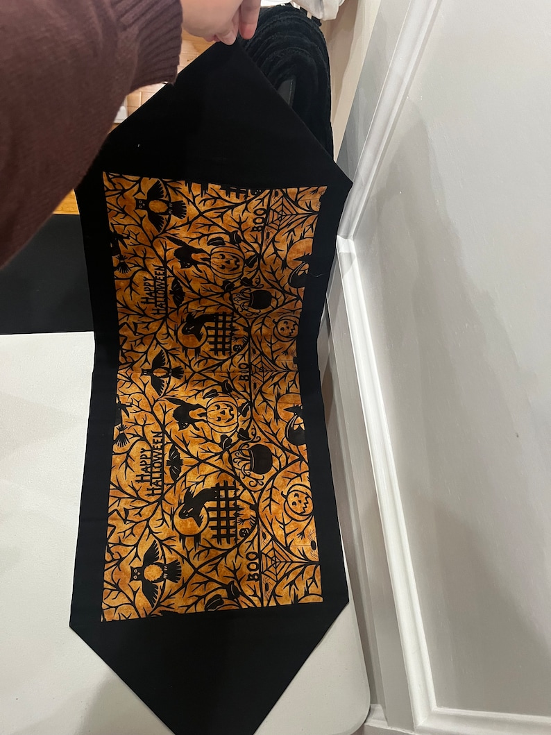 May include: Black table runner with a gold and black Halloween design. The design features owls, pumpkins, bats, and the words "Happy Halloween" and "Boo".