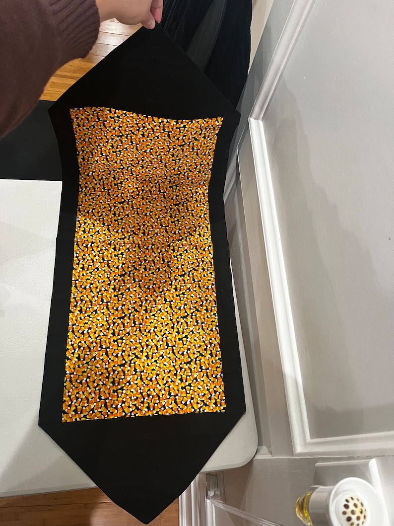 May include: Black table runner with a pattern of orange and white candy corn. The runner has a pointed end.