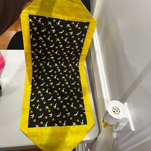 May include: A yellow table runner with a black and white pattern of flying birds and yellow circles. The runner has a yellow border.
