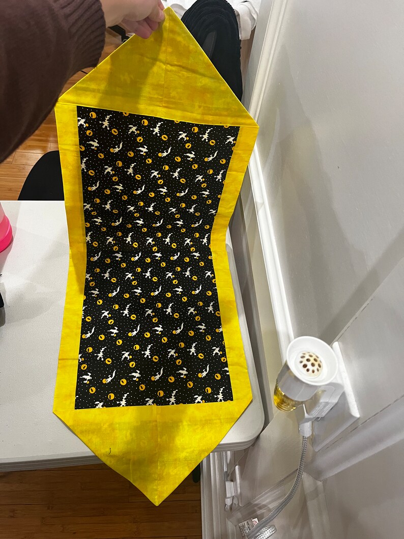 May include: A yellow table runner with a black and white pattern of flying birds and yellow circles. The runner has a yellow border.