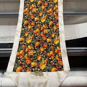 May include: A table runner with a black background and a pattern of orange pumpkins and green leaves. The runner has a beige border with a wheat design.