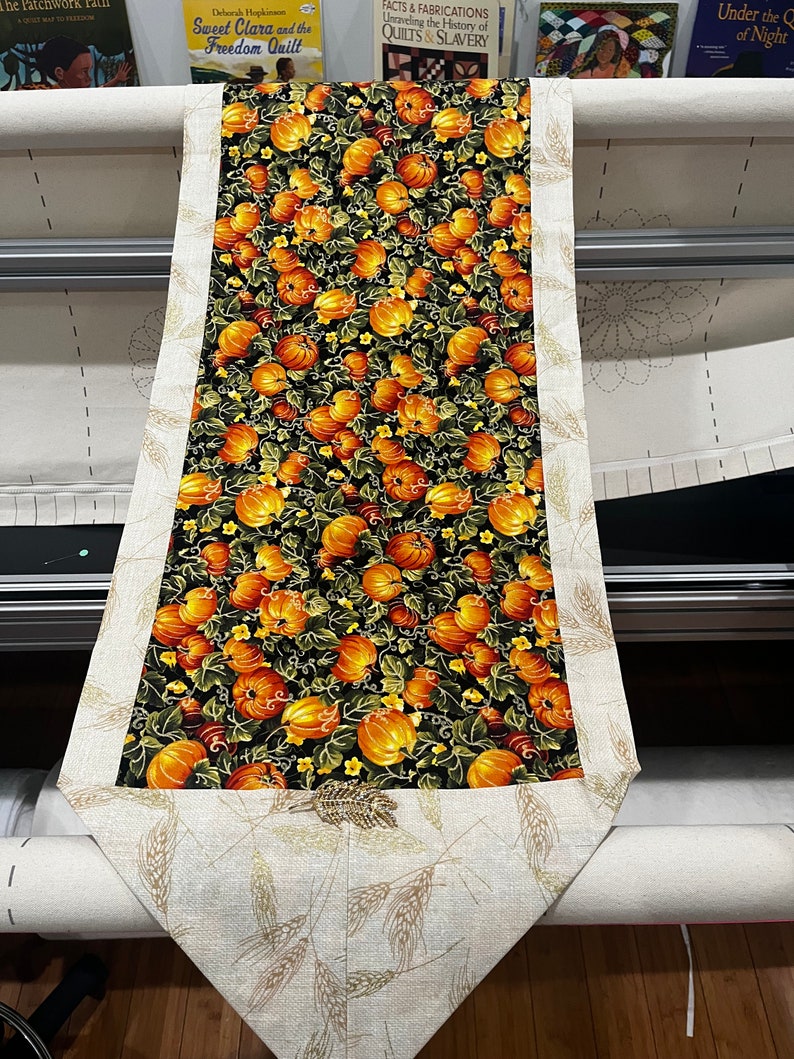 May include: A table runner with a black background and a pattern of orange pumpkins and green leaves. The runner has a beige border with a wheat design.