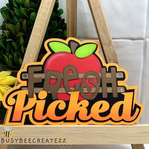 May include: A red apple with green leaves and a brown wooden sign that says "Fresh Picked" in orange and black letters.