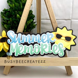 May include: A decorative sign featuring the words "Summer Memories" in blue and green, with a yellow sun wearing sunglasses. The sign is displayed on a small wooden easel, with the text "BUSYBEECREATEZZ" at the bottom.