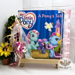 Könnte beinhalten: Ein My Little Pony A Pony's Tale Buchcover mit zwei Ponys und einer Schatztruhe, verziert mit einer seitlichen Quaste aus rosa und weißen Bändern, Tüll und einem kleinen Pony-Anhänger, präsentiert auf einem kleinen hölzernen Staffeleiständer.
