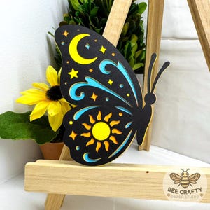 May include: A layered paper butterfly cutout featuring a black top layer with cutouts revealing yellow and light blue layers underneath. The design includes a yellow crescent moon, a yellow sun, and various star shapes. A small bee logo is visible.