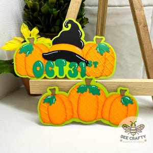 May include: Two layered paper craft decorations featuring orange pumpkins with a grid texture, green stems, and bright green leaves. The top piece includes a black witch hat and the green text OCT 31ST. Both items have a vibrant lime green border.