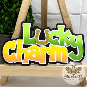 May include: A layered paper cutout with the words "Lucky Charm" in gradient yellow and green, with a gold glitter horseshoe. The cutout is on a black background and displayed on a small wooden easel. The Bee Crafty Paper Studio logo is visible.
