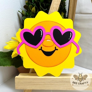 May include: A die-cut paper sun decoration featuring a bright yellow jagged border, an orange circular face, a smiling mouth, and pink heart-shaped sunglasses with black lenses. The design includes a small Bee Crafty Paper Studio logo.