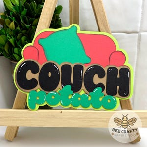 May include: A colourful paper art piece depicting a red sofa with a green cushion. The words "COUCH potato" are in black bubble letters with a green outline. The piece is displayed on a small wooden easel, with a "Bee Crafty Paper Studio" logo.
