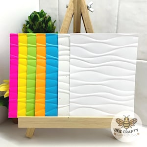 Peut inclure: Présentation de papiers texturés aux couleurs vives : rose, jaune, vert, orange et bleu, ainsi qu'un papier blanc à motif ondulé. Les échantillons sont exposés sur un petit chevalet en bois, mettant en valeur les textures et les couleurs.