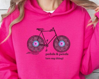 Op de afbeelding: Een roze hoodie met een zwart-witte illustratie van een fiets met bloemmotieven op de wielen. De tekst "pedals & petals (are my thing)" staat onder de fiets gedrukt.