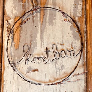 Könnte beinhalten: Ein kreisförmiger Drahtkranz mit dem Wort "kostbar" in Schreibschrift. Der Kranz ist mit kleinen Zweigen getrockneter Blumen geschmückt und vor einem verwitterten Holzgrund platziert.