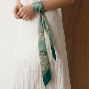 May include: A light green silk scarf tied around a wrist. The scarf features a floral pattern with a bird design and dark green borders. The scarf is made of a soft, flowing material.