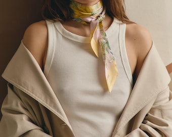 May include: A white ribbed tank top worn under a beige trench coat, paired with a silk neck scarf featuring a yellow, pink, and green floral botanical print. The scarf is tied in a simple knot at the front, resting against the white ribbed fabric.