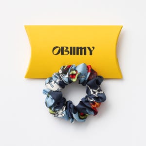 May include: A colorful scrunchie with a blue, red, and white pattern sits in front of a yellow box. The scrunchie has a mix of floral and abstract designs. The box has the word "OBIIMY" printed on it in black.