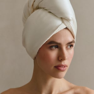May include: A hair towel wrap in a solid off-white color. The exterior of the wrap features a smooth, satin-like finish, while the interior lining is composed of a textured, absorbent terry cloth fabric. The wrap is secured in a twisted style.