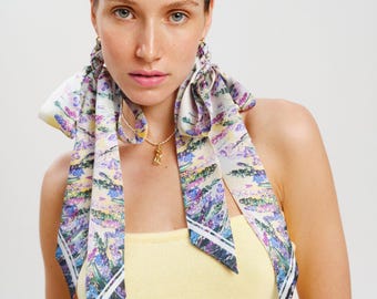 May include: A long, narrow silk scarf featuring a vibrant floral print with shades of purple, green, and yellow on a cream background. The scarf is tied around the hair, with two long ends draping down the sides, and includes a small gold pendant necklace.